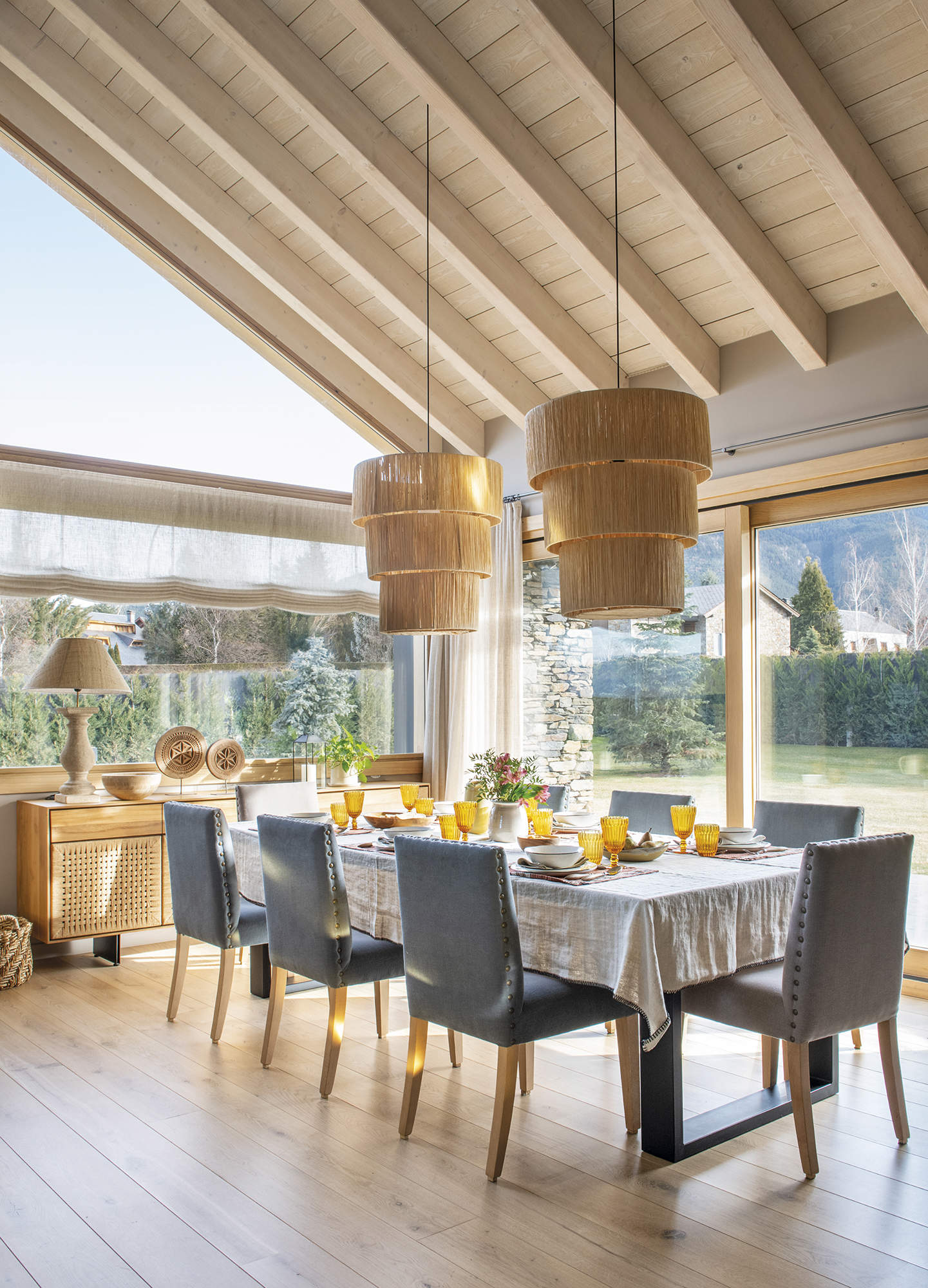 comedor, mesa con mantel con sillas tapizadas color gris, lámparas de techo de fibra, aparador con lámpara de mesa, suelo de madera, techo con vigas